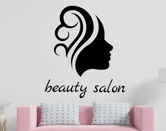 Può includere: Silhouette nera di un profilo femminile con capelli stilizzati, sopra le parole "beauty salon". Il design è su un muro bianco sopra un divano rosa con due cuscini. Sono visibili anche una lampada e un tavolino.