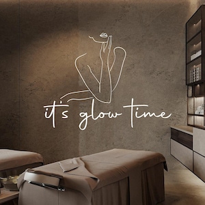 Può includere: Una sala per trattamenti spa con due lettini da massaggio, una parete bianca con un disegno a linea di una donna e il testo "it's glow time", e un armadio con ripiani.