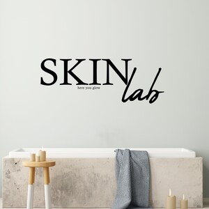 May include: A white bathtub with a grey towel hanging over the side. The wall behind the tub has a black and white sign that reads "SKIN lab here you glow".