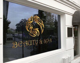 Può includere: Una vetrina di un negozio con un logo dorato che presenta un design floreale stilizzato e il testo "BEAUTY & SPA".