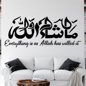 May include: A white wall with a black Arabic calligraphy design and the text "Everything is as Allah has willed it". The design is in a living room setting with a white couch and pillows.