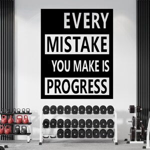 May include: Black and white motivational quote on a wall in a gym setting. The quote reads "Every mistake you make is progress". The quote is displayed on a black background with white text. The gym has a weight rack with dumbbells and a weight bench.