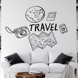 Op de afbeelding: Een witte bank met zwarte kussens in een woonkamer met een witte muur waarop een zwart-witte illustratie van een wereldbol, een kaart, een camera, een paspoort en het woord "TRAVEL" te zien is.