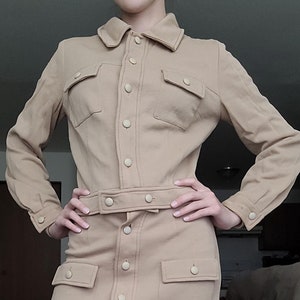 Op de afbeelding: Een beige, langarmige knoopjurk met een riem en grote zakken. De jurk heeft een kraag en een aansluitende pasvorm.