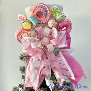 May include: A decorative tree topper featuring a large pink and white striped bow with glittery accents. The arrangement is adorned with various faux candy elements including pink donuts, colorful cake pops, and peppermint swirls in shades of pink, white, and green. The base is a white fabric structure.