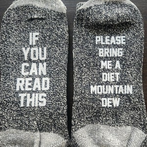 Puede incluir: Un par de calcetines grises y negros con texto blanco. Un calcetín dice "IF YOU CAN READ THIS" y el otro "PLEASE BRING ME A DIET MOUNTAIN DEW". Los calcetines tienen puño y puntera grises.