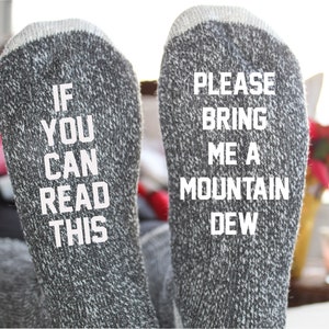 Puede incluir: Un par de calcetines grises con texto blanco. Un calcetín dice "Si puedes leer esto" y el otro calcetín dice "Por favor, tráeme una Mountain Dew."