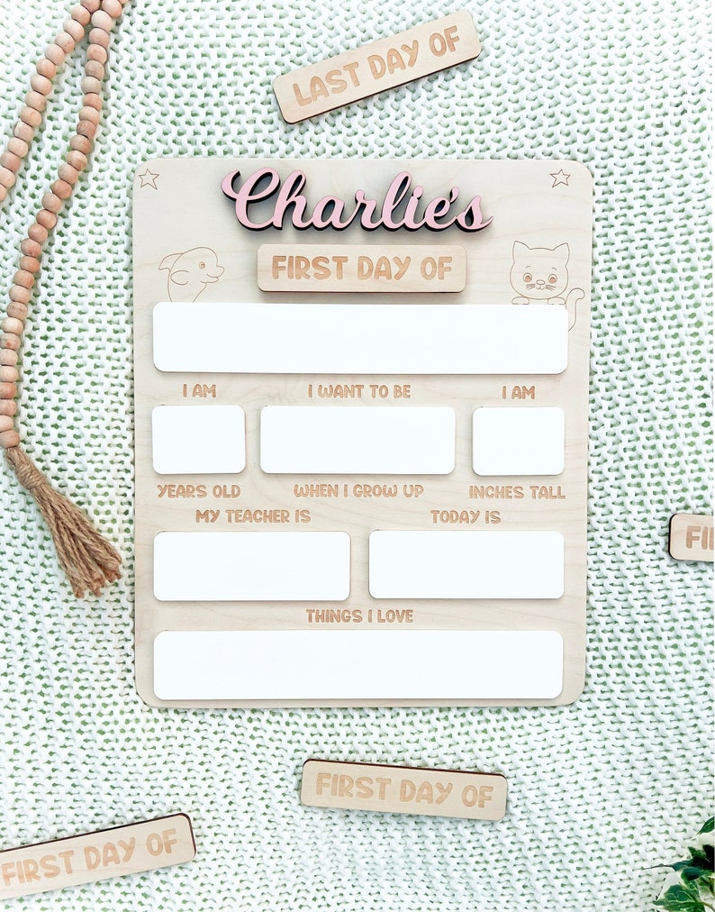 May include: A wooden sign with the name "Charlie's" in pink lettering and the words "First Day Of" on a separate line. The sign has spaces for writing information about the child's age, height, teacher, and things they love. There are also spaces for writing about their hopes and dreams. The sign is decorated with a star, a dolphin, and a cat.