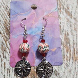 May include: A pair of dangle earrings with a silver compass charm and a white and green patterned bead with red accents.