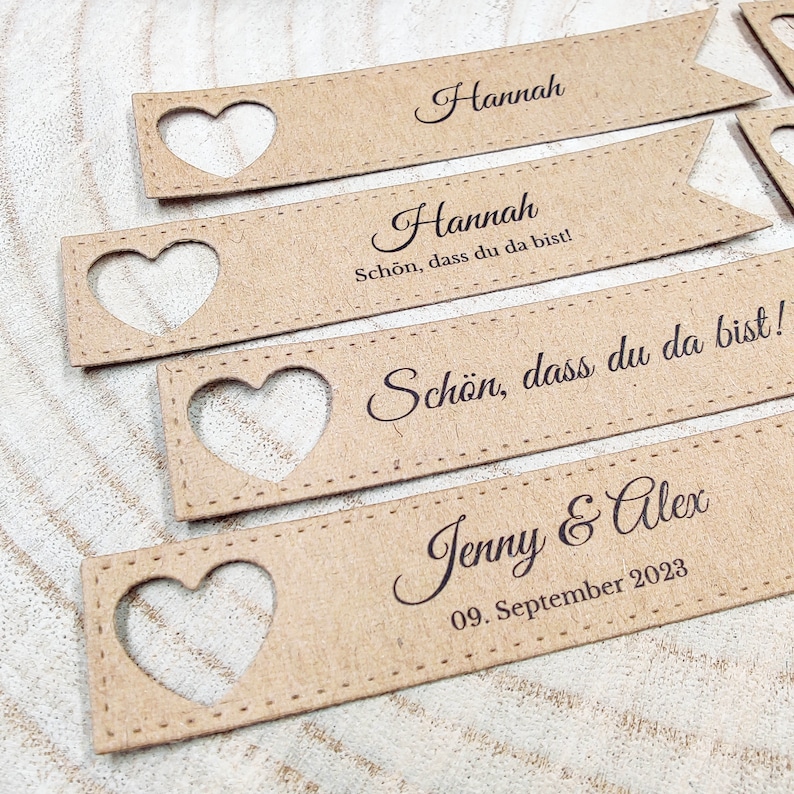 May include: Set of brown paper place cards with a heart cut-out. The cards have the text "Hannah" and "Sch&ouml;n, dass du da bist!" printed on them. One card has the text "Jenny & Alex" and "09. September 2023" printed on it.