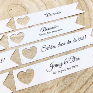 May include: White paper place cards with a heart-shaped cutout. The text on the cards reads "Alexander" and "Sch&ouml;n, dass du da bist!"  The last card reads "Jenny & Alex 09. September 2023".