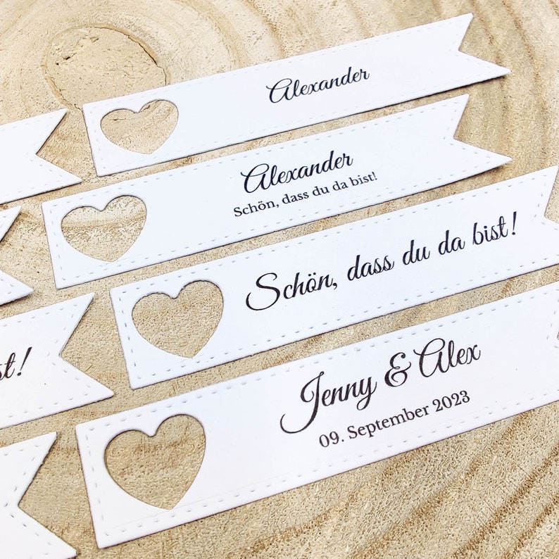 May include: White paper place cards with a heart-shaped cutout. The text on the cards reads "Alexander" and "Sch&ouml;n, dass du da bist!"  The last card reads "Jenny & Alex 09. September 2023".