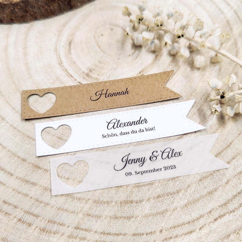 May include: Three brown and white paper place cards with a heart cut-out. The top card reads "Hannah". The middle card reads "Alexander" and "Sch&ouml;n, dass du da bist!" The bottom card reads "Jenny & Alex" and "09. September 2023".