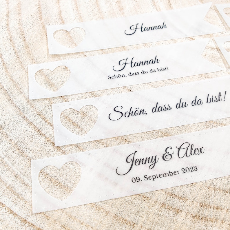 May include: Four white place cards with a heart-shaped cutout. The top two cards read "Hannah" and "Hannah, Sch&ouml;n, dass du da bist!" The bottom two cards read "Sch&ouml;n, dass du da bist!" and "Jenny & Alex, 09. September 2023".