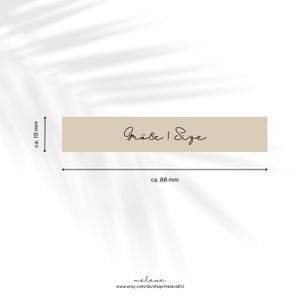 May include: A beige label with the text "Gr&ouml;&szlig;e | Size" in black cursive writing. The label is approximately 8.8 cm wide and 1.9 cm tall.