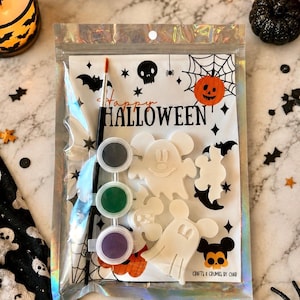 May include: A Halloween craft kit in a holographic pouch containing three white ceramic shapes resembling ghosts with mouse ears, a small black paintbrush, and three small circular paint pots in black, green, and purple. The packaging features the text Happy HALLOWEEN and CRAFTS & CRUMBS BY CHAR.