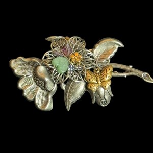 May include: Silver tone brooch with a flower design featuring a green stone, a butterfly, and other colorful accents.