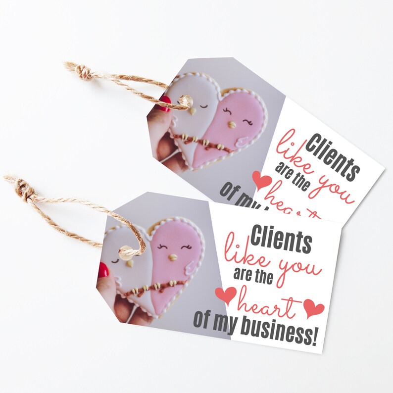 Puede incluir: Dos galletas con forma de coraz&oacute;n decoradas con glaseado rosa y blanco y ojos pintados. Las galletas est&aacute;n sostenidas por una mano y el texto "Clients like you are the heart of my business!" est&aacute; escrito en la etiqueta.