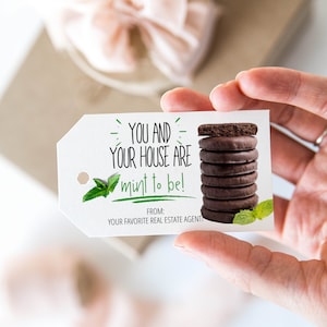 May include: A white gift tag with green text that reads "YOU AND YOUR HOUSE ARE mint to be!"  The tag features a stack of chocolate mint cookies and a green leaf design. The text "FROM: YOUR FAVORITE REAL ESTATE AGENT!" is printed at the bottom of the tag.