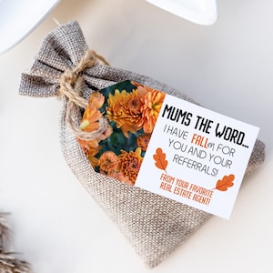 May include: A white gift tag with the text "MUMS THE WORD... I HAVE FALLEN FOR YOU AND YOUR REFERRALS! FROM YOUR FAVORITE REAL ESTATE AGENT!"  The tag is decorated with orange and brown fall leaves. The tag is attached to a small burlap bag filled with orange mums.