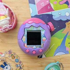 Könnte beinhalten: Ein lila-rosa Handheld-Digital-Haustierspiel mit schwarzem Bildschirm und rosa Knöpfen. Weitere Artikel sind eine transparente rosa Hülle mit einer weißen Figur, ein grüner und blauer Schlüsselanhänger und ein transparenter Schlüsselanhänger mit farbenfrohen Grafiken.