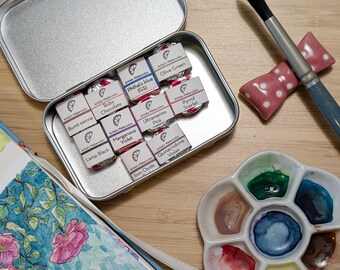 May include: A metal tin filled with 12 watercolor paint pans. The pans are labeled with the names of the colors, including Burnt Sienna, Lamp Black, Ruby Chocolate, Phthalo Blue (GS), Ultramarine Pink, Manganese Violet, Iron Oxide, Quinacridone Violet, Pyrrol Scarlet, and Olive Green.