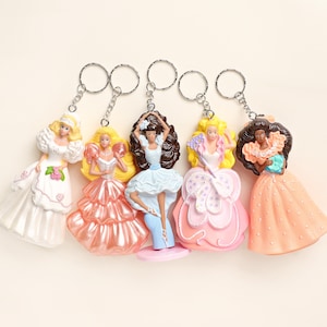 Puede incluir: Cinco llaveros de muñecas con diferentes vestidos y peinados. Las muñecas tienen cabello rubio, castaño y negro. Los vestidos son en tonos de blanco, rosa y melocotón. Cada llavero tiene un anillo plateado.