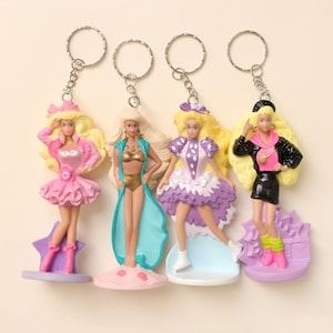 Puede incluir: Cuatro llaveros de muñecas vintage. Cada muñeca tiene cabello rubio y viste un atuendo diferente. Los atuendos incluyen un vestido rosa, un traje de baño dorado con una cubierta azul, un vestido morado y una chaqueta negra con top y falda rosas.