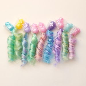 May include: A collection of colorful hair curlers in various pastel shades. Each curler features a small, animal-shaped clip at the top, with the curls themselves ranging from light blue to pink and purple. The curlers are arranged on a light-colored surface.