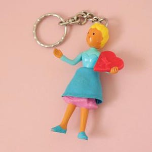 May include: A keychain featuring a small figurine with yellow hair, an orange face, and a blue dress over a pink skirt. The figure holds a red heart-shaped object in one hand and is attached to a silver-colored metal split ring by a short chain.