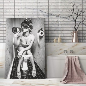 May include: Black and white photo of a woman sitting on a toilet in a bathroom, drinking from a bottle. The woman is wearing sunglasses and has her hair in rollers. The bathroom is tiled and has a toilet paper holder.