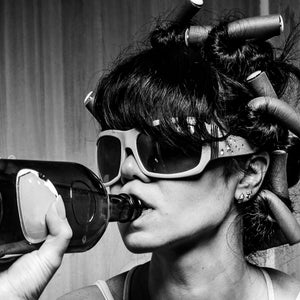 May include: A woman with dark hair and sunglasses is drinking from a glass bottle. Her hair is styled with rollers.