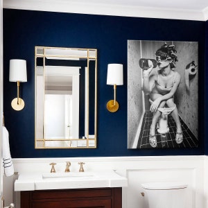 May include: A bathroom with a dark blue wall, a white vanity with a gold faucet, a gold framed mirror, and a black and white print of a woman sitting on a toilet.
