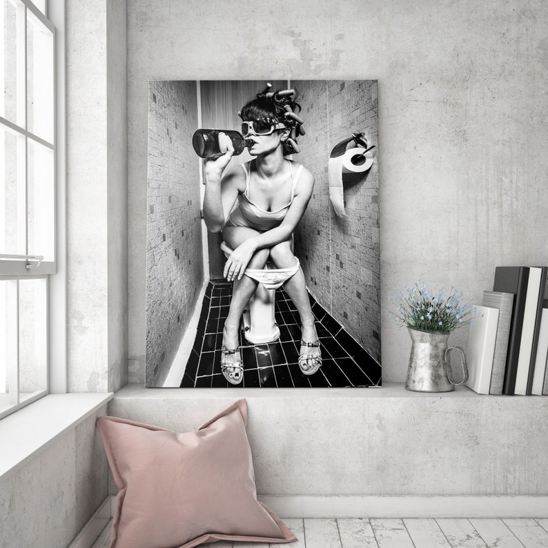 May include: Black and white photo of a person sitting on a toilet in a bathroom, drinking from a bottle. The person is wearing sunglasses and has their hair up in a bun. There is a roll of toilet paper on the wall behind them.