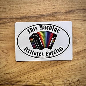 Peut inclure: Un autocollant rectangulaire avec un design ovale. Le design présente un accordéon noir avec un soufflet aux couleurs de l'arc-en-ciel. Le texte se lit "This Machine Irritates Fascists" et "AccordionRevolution.com".