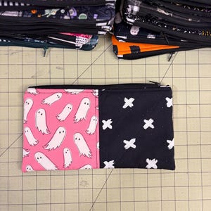 May include: A rectangular, zippered pouch. One side is pink with white ghost illustrations, the other black with white cross patterns. Other pouches with Halloween patterns are visible in the background.