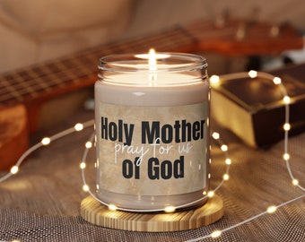 May include: A lit candle in a clear glass jar with the text "Holy Mother pray for us of God" on a tan label. The candle sits on a small wooden coaster, with string lights and a guitar in the background.