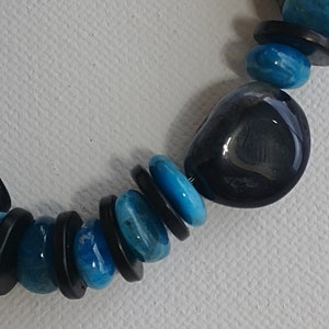 May include: A necklace featuring a repeating pattern of smooth, round, bright turquoise blue beads and thin, flat, circular black spacer beads. The design includes one larger, irregular, polished dark charcoal grey stone bead among the smaller beads.