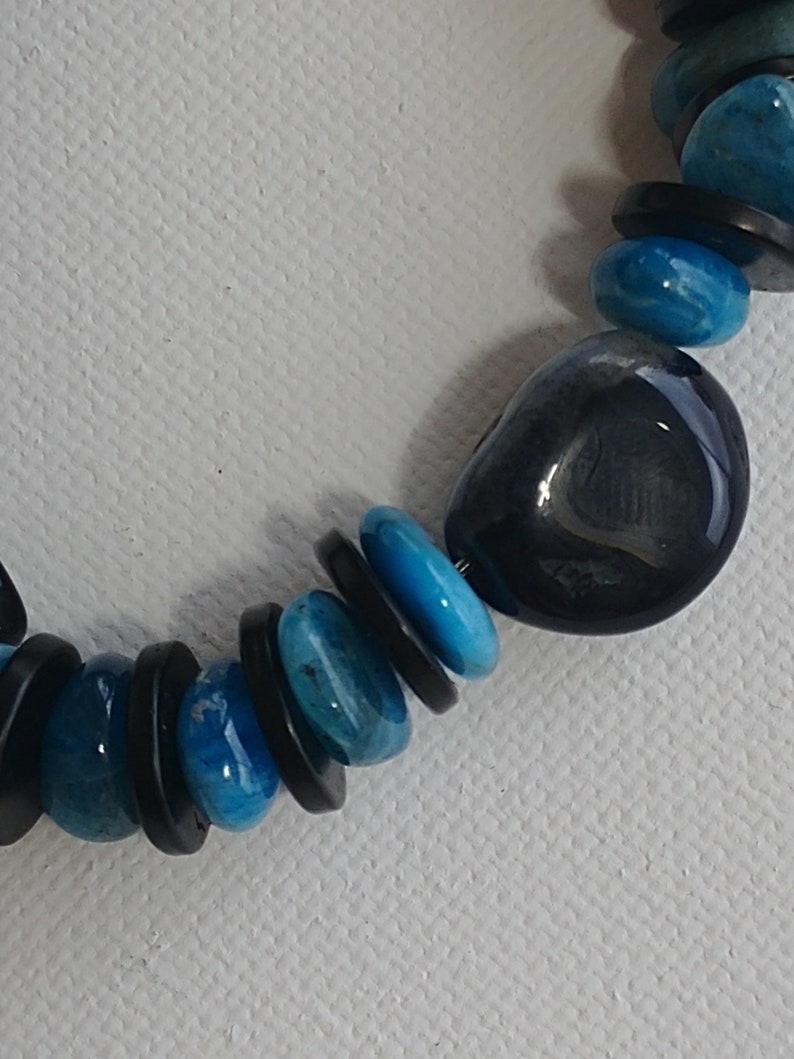 May include: A necklace featuring a repeating pattern of smooth, round, bright turquoise blue beads and thin, flat, circular black spacer beads. The design includes one larger, irregular, polished dark charcoal grey stone bead among the smaller beads.