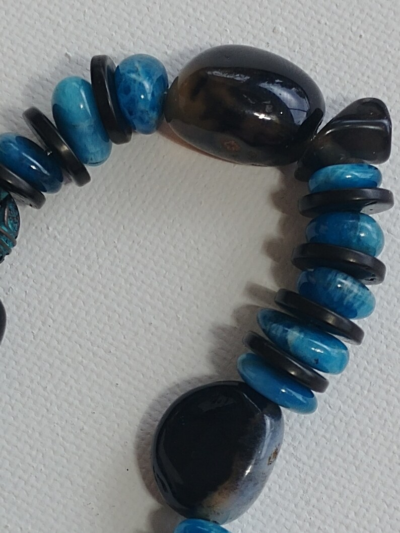 May include: A bracelet featuring alternating smooth, round, bright blue beads and thin, flat, dark brown disc-shaped spacers. Two large, polished, dark brown, irregular oval-shaped stones are positioned at intervals along the beaded strand.