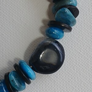 May include: A beaded necklace featuring alternating round, polished blue stones and thin, flat black circular spacers. A larger, smooth, dark charcoal-coloured stone with a lighter grey centre is positioned as a focal point among the blue and black beads.