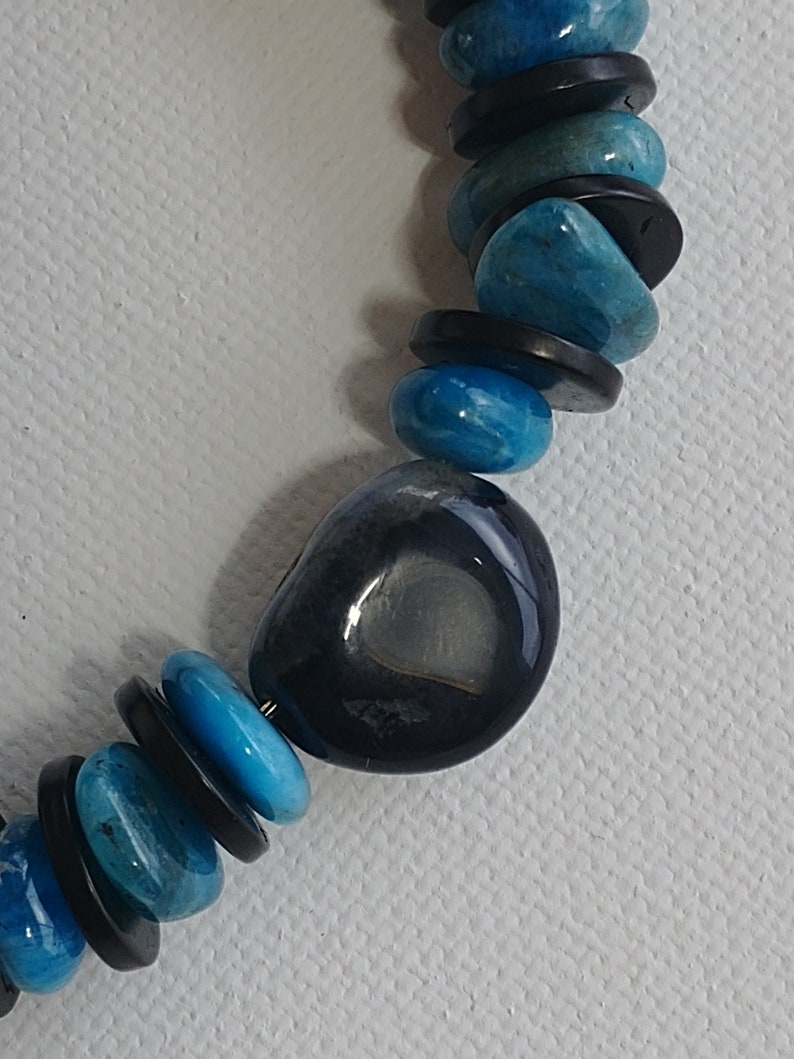 May include: A beaded necklace featuring alternating round, polished blue stones and thin, flat black circular spacers. A larger, smooth, dark charcoal-coloured stone with a lighter grey centre is positioned as a focal point among the blue and black beads.