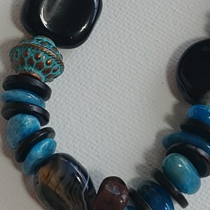 May include: A beaded necklace featuring a mix of smooth black square beads, round turquoise-coloured stones, thin black disc spacers, and a textured copper-toned bead with a teal patina. The strand also includes a polished brown and grey banded agate stone.