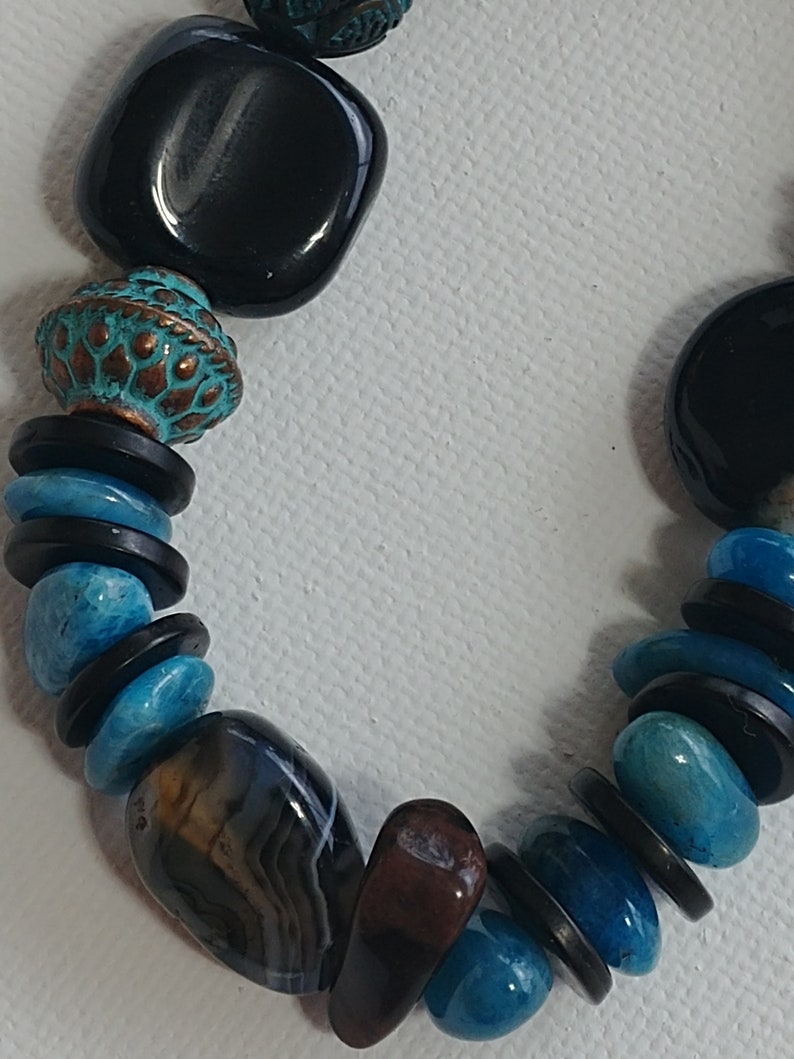 May include: A beaded necklace featuring a mix of smooth black square beads, round turquoise-coloured stones, thin black disc spacers, and a textured copper-toned bead with a teal patina. The strand also includes a polished brown and grey banded agate stone.