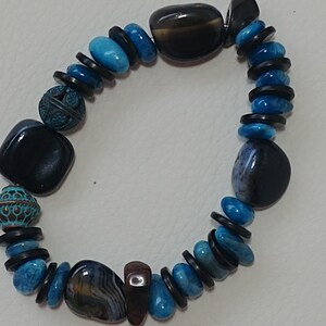 May include: A beaded bracelet featuring a mix of polished black and brown agate stones, vibrant blue turquoise-coloured beads, and small black disc spacers. The design includes two round, textured metal beads with a dark teal patina finish.
