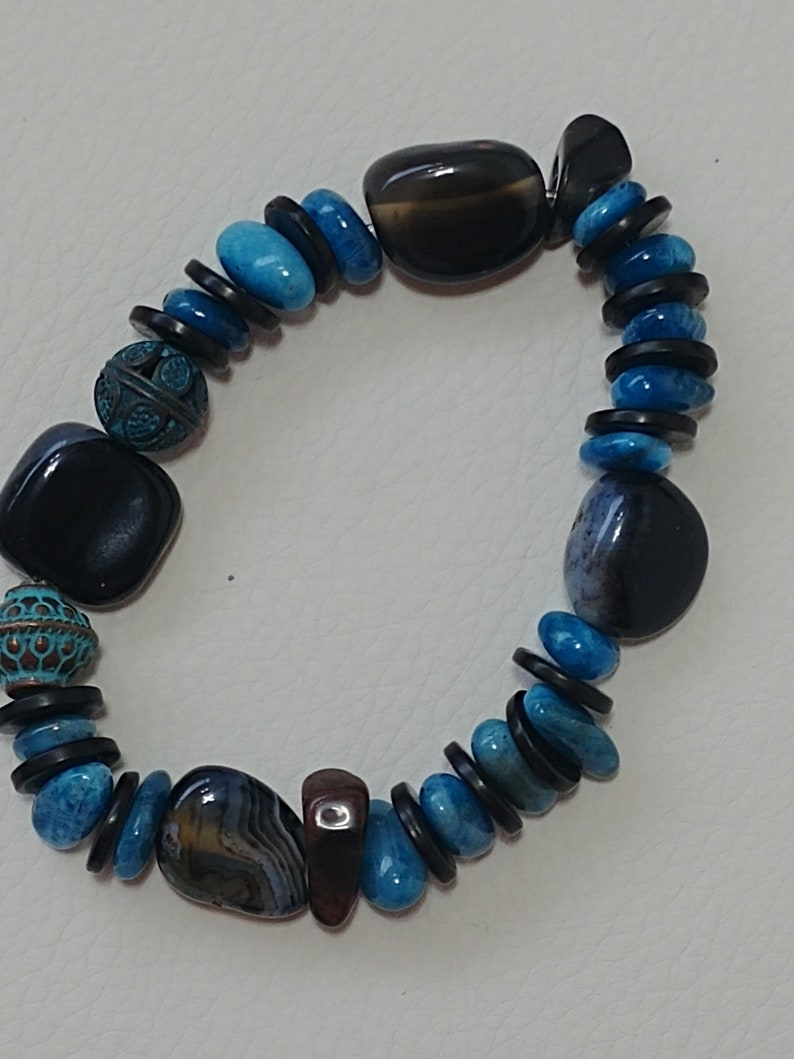 May include: A beaded bracelet featuring a mix of polished black and brown agate stones, vibrant blue turquoise-coloured beads, and small black disc spacers. The design includes two round, textured metal beads with a dark teal patina finish.