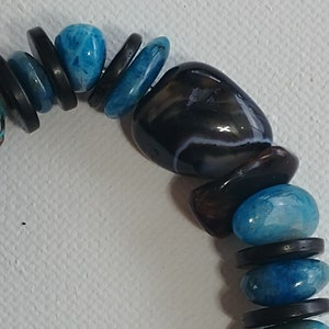 May include: A beaded bracelet featuring alternating smooth, polished blue stones and flat, circular black discs. The design includes a larger, dark, rounded stone with subtle white banding, creating a contrast between the vibrant blue and deep black elements.