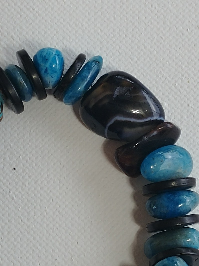May include: A beaded bracelet featuring alternating smooth, polished blue stones and flat, circular black discs. The design includes a larger, dark, rounded stone with subtle white banding, creating a contrast between the vibrant blue and deep black elements.