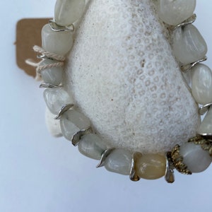 May include: A bracelet composed of smooth, tumbled, translucent white and pale yellow stones. The stones are separated by thin, curved silver-toned metal spacers. One end of the bracelet features a decorative, textured gold-toned metal cap.