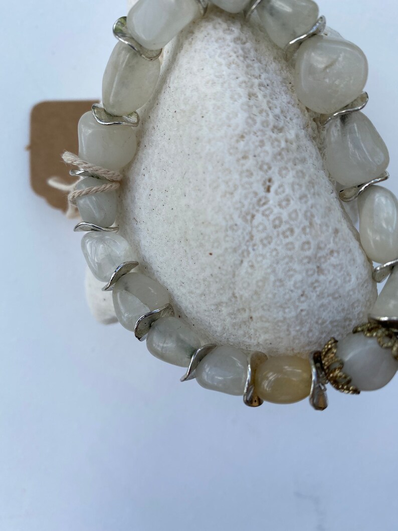 May include: A bracelet composed of smooth, tumbled, translucent white and pale yellow stones. The stones are separated by thin, curved silver-toned metal spacers. One end of the bracelet features a decorative, textured gold-toned metal cap.